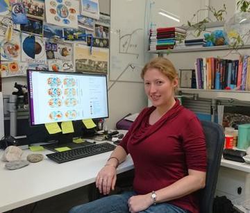 A photograph of Paula sitting in her office. She is sitting on a black chair next to a white desk with a computer and some rocks on it. Behind her are some white bookshelves. There are lots of images of the Earth on the wall.