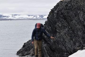 An image of Ross walking across some dark grey rocks on the edge of a lake. There are snow mountains in the background. He is wearing a blue coat and an orange baseball cap.