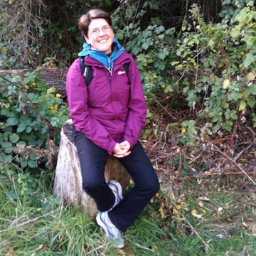 Photograph of Elizabeth Crowley. She is wearing a purple jacket and black trousers, has short hair, and is sat on a tree stump smiling at the camera.