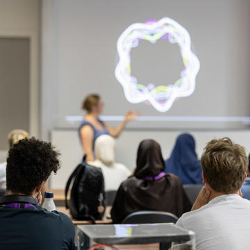 Photograph of a lecturer stood at the front of a class pointing to slides on the wall behind, which display vibrations in the Earth