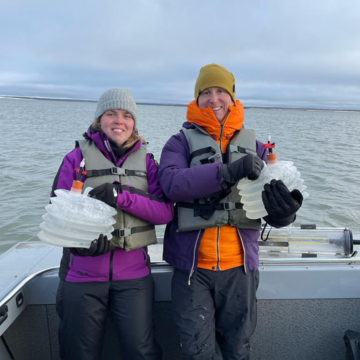 Dr Madeleine Stow and Professor Bob Hilton conducting geochemical fieldwork in Canada. They are standing on a boat wearing thick coats and life vests, and holding large containers of water.