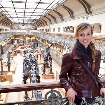 Photograph of Erin Saupe standing on a balcony in a museum with exhibits behind her