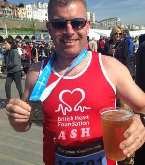 Photograph of Ashleigh Hewson. He is smiling at the camera wearing a red British Heart Foundation running vest and holding a medal and a drink.