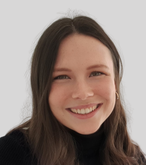 Photograph of Charlie smiling at the camera. She is wearing a black polo neck top and has long wavy brown hair.