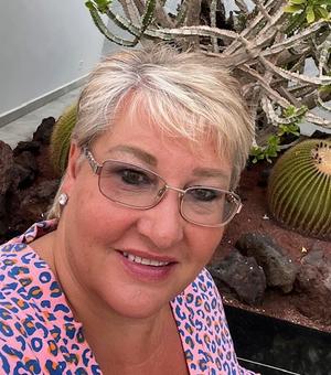 Photograph of Jeanette Stimpson. She is wearing glasses and a pink patterned top and smiling at the camera.